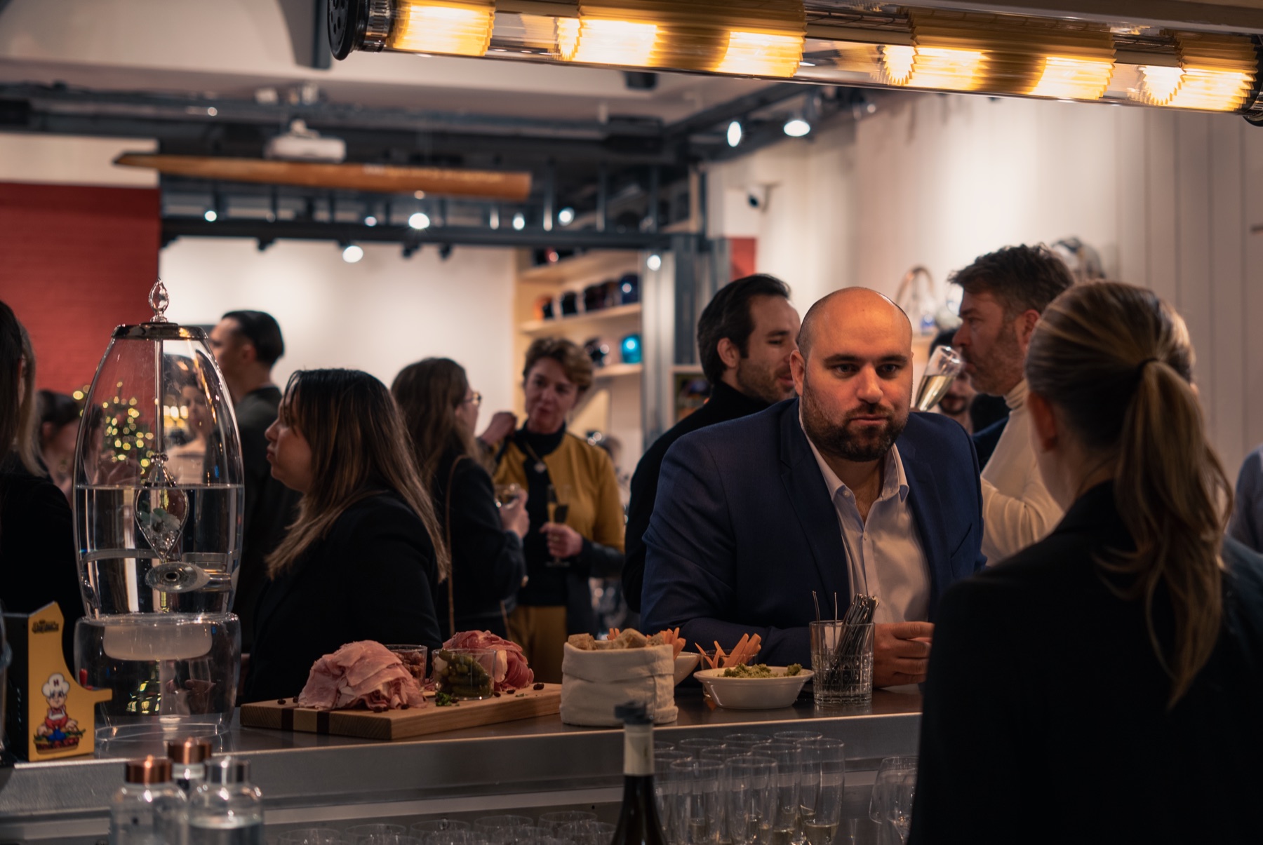 Soirée d'entreprise au bar de La Fabrique Générale · invités en discussion, planche de charcuterie et frites, fontaine à eau cristalline, lumière chaude